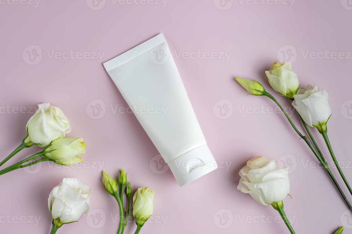 Blank plastic white tube for cream, mask, shampoo or toothpaste on pink background among eustoma flowers. Spring presentation of cosmetics package. Mockup photo