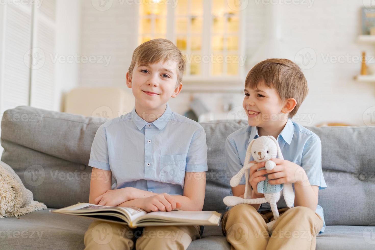 Two boys sitting on couch are reading book. Older brother reads interesting photo