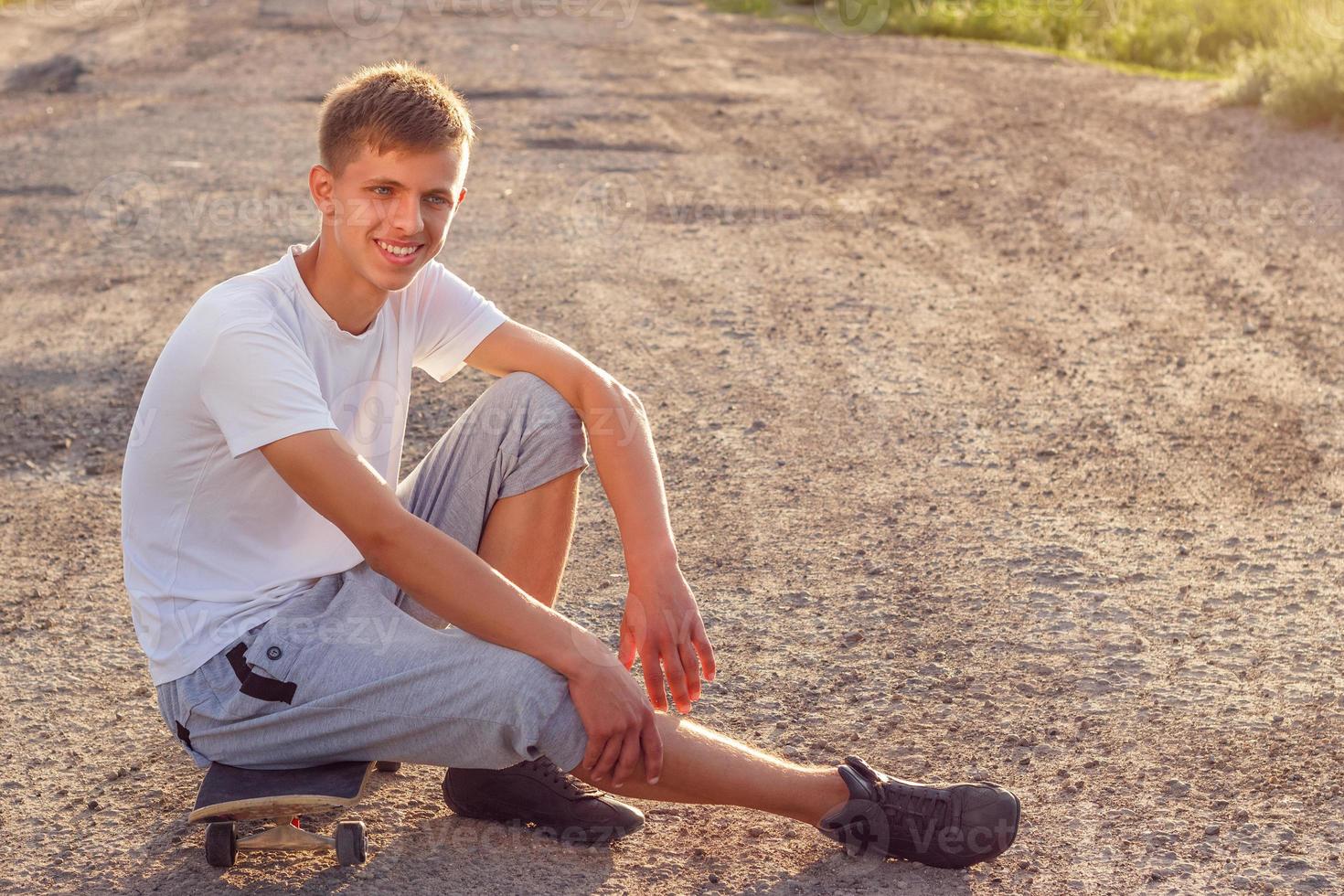 chico alegre sentado en una patineta en la carretera en un día soleado foto