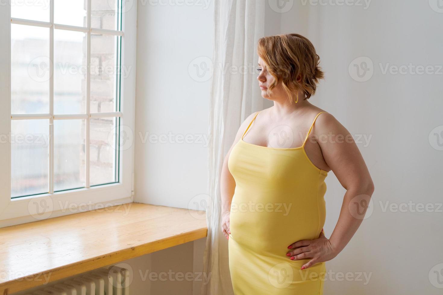 Adult plus size woman in tight-fitting yellow dress, makeup standing by window photo