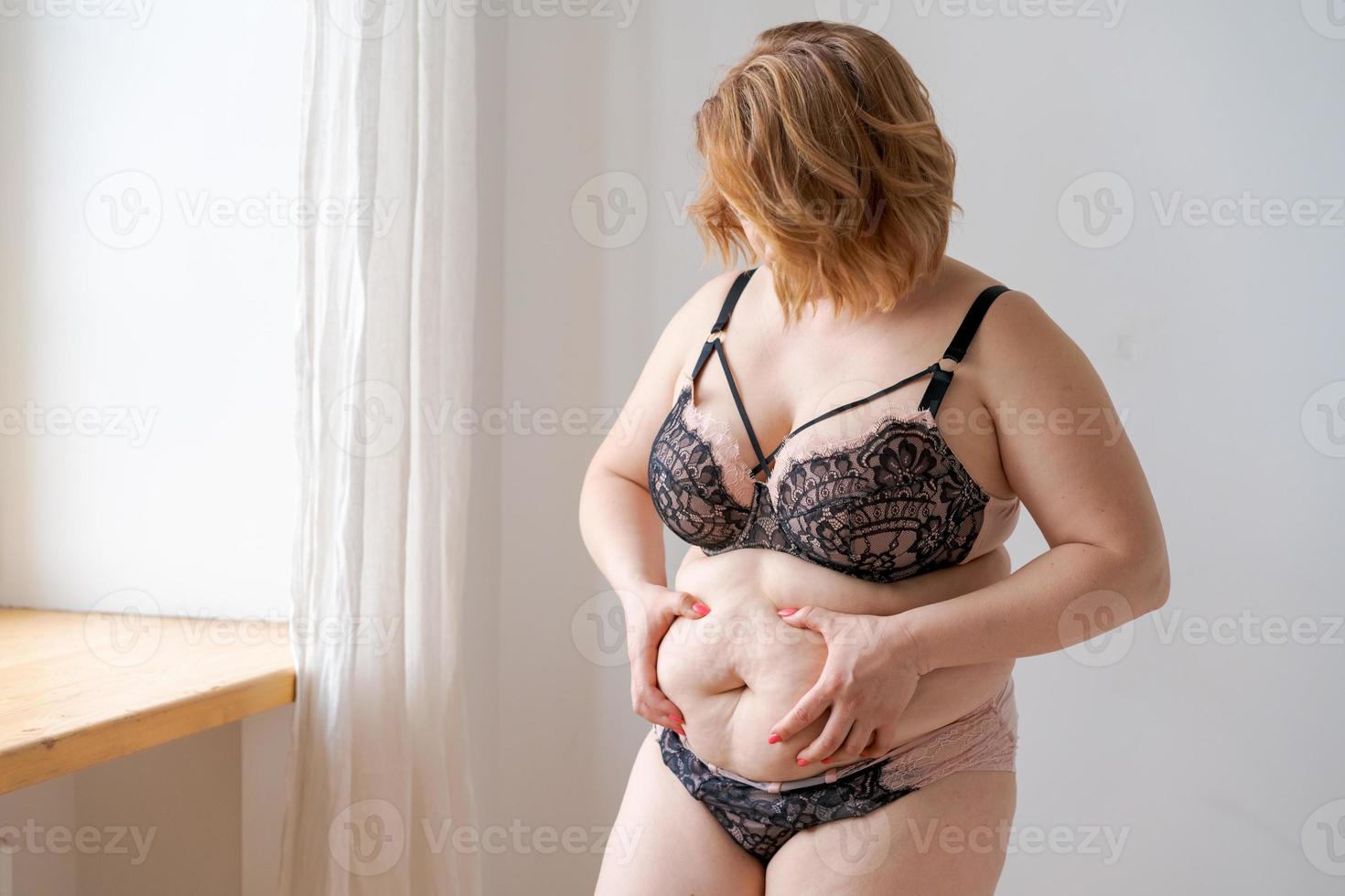 Obese plump caucasian young woman, girl holding her belly by hand, shows excess photo