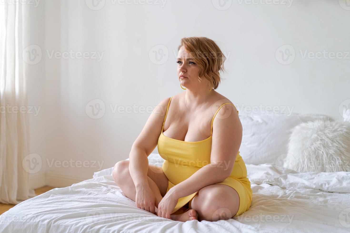 Plus-size model in yellow slinky dress sits on bed beside her bedroom photo