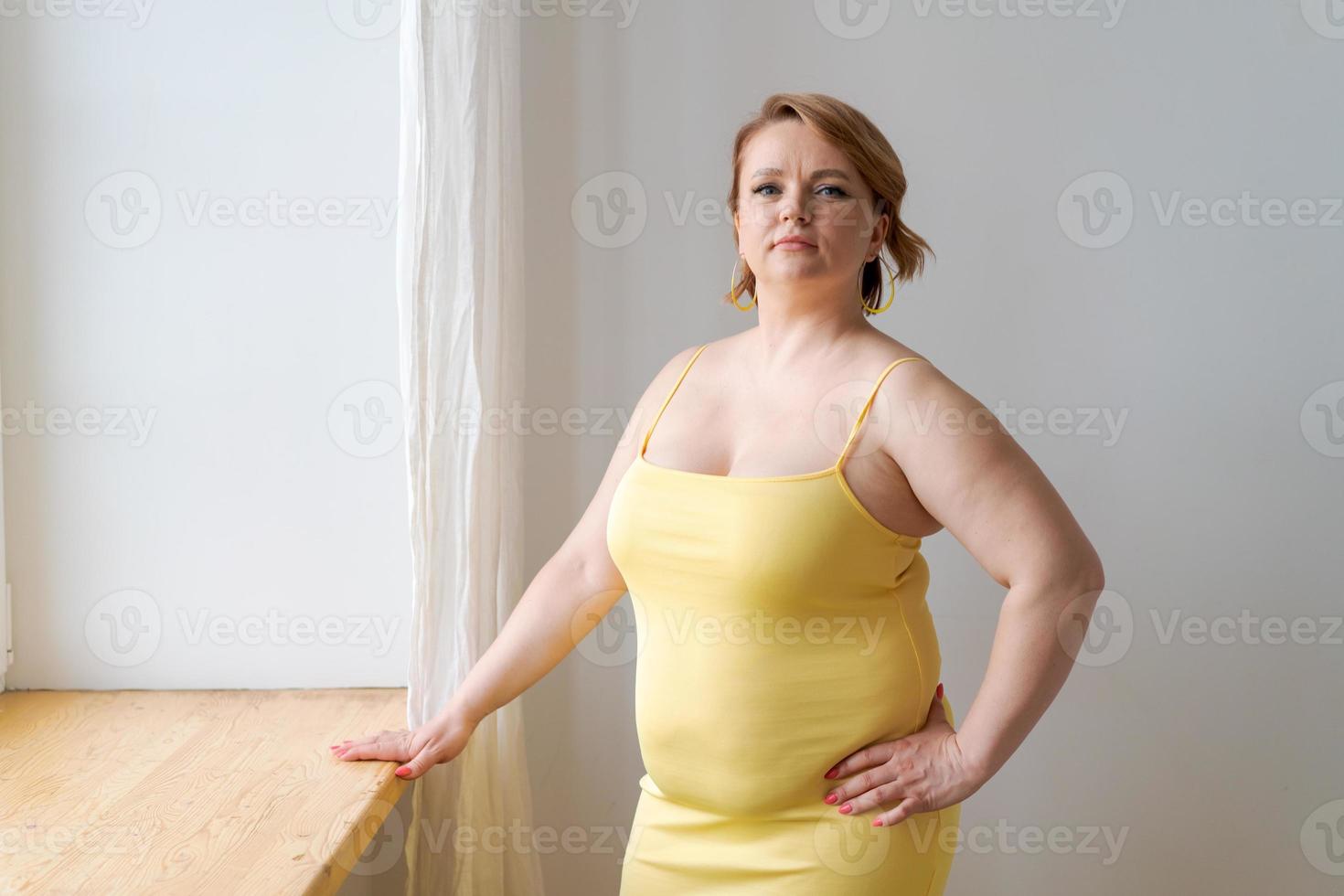 mujer adulta de talla grande con vestido amarillo ajustado, maquillaje de pie junto a la ventana foto