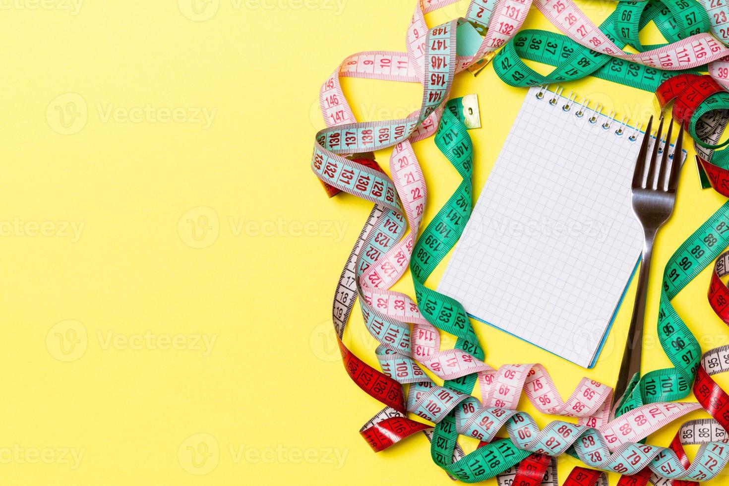 Fitness concept with empty space for your design on yellow background. Top view of notepad and fork surrounded with colorful measure tapes photo