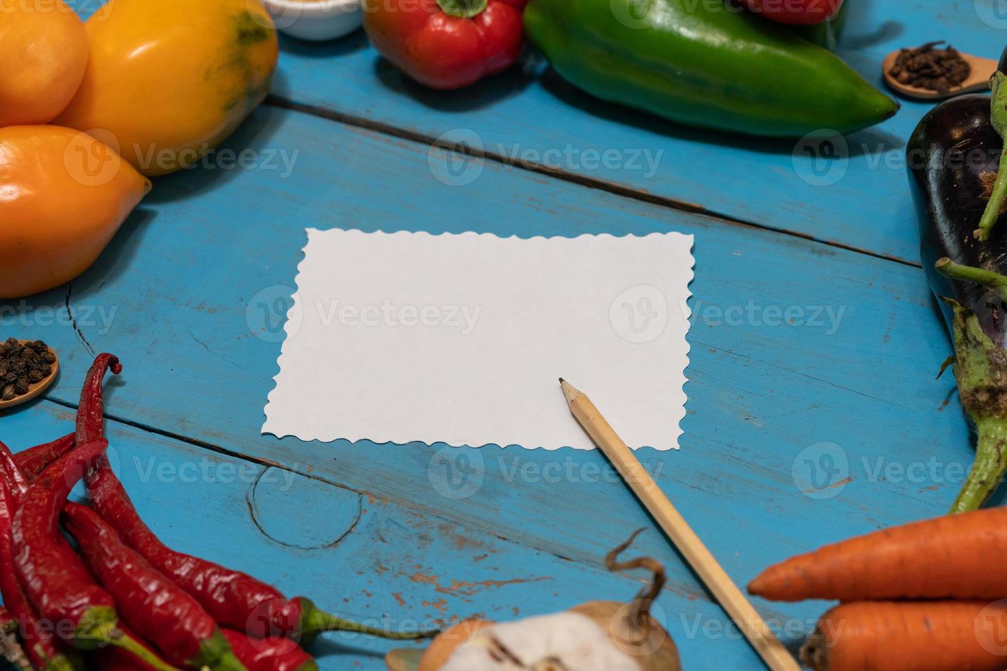 Vegetables are laid out around a sheet of paper and a pencil. Empty space for text. Vegetables, empty blank for recipe on a blue background. photo