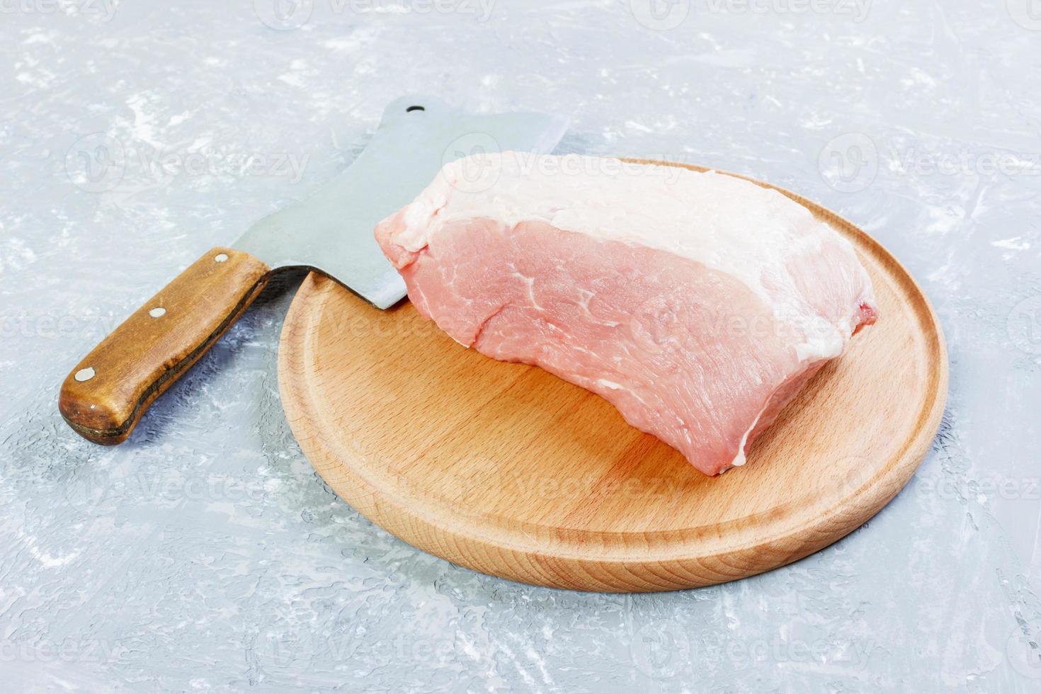 pieza grande de carne de cerdo fresca en una tabla redonda de madera con hacha de cocina. vista superior sobre fondo de hormigón gris foto