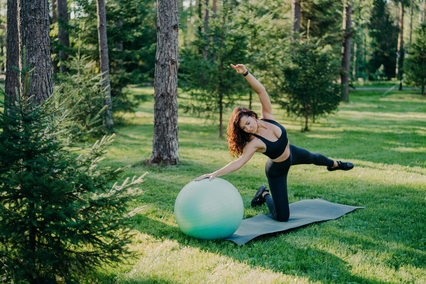Fitness, sport and healthy lifestyle concept. Outdoor shot of slim brunette European woman does pilates and balance exercise with fit ball tilts aside poses in green forest on grass at karemat. photo