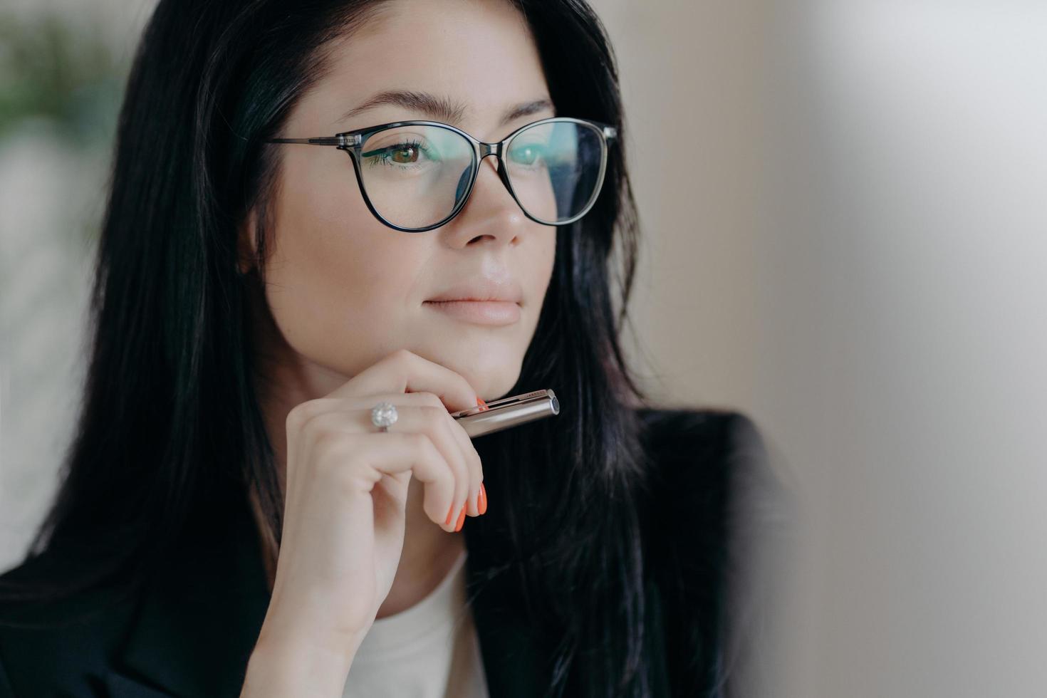 Serious woman author works on new publication, concentrated in display of computer, keeps pen in hand, wears spectacles for eye correction. Ambitious successful female entrepreneur manages business photo