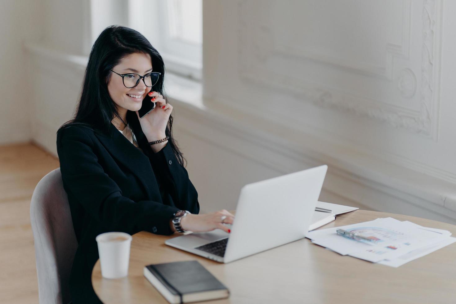 Beautiful female executive makes remote job, talks via mobile phone and checks information in laptop computer poses at workplace surrounded by coffee paper documents notepad has telephone conversation photo