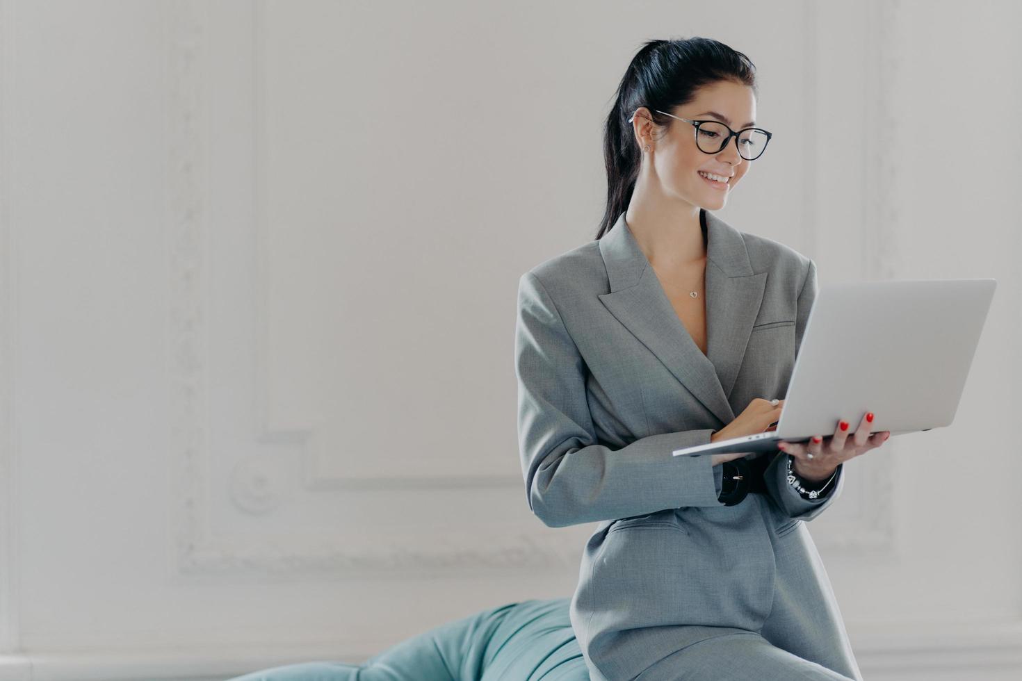 Happy female entrepreneur types on laptop computer, works in modern workspace in business center, dressed in formal outfit, surfes internet, watches training webinar, chats online with partners photo