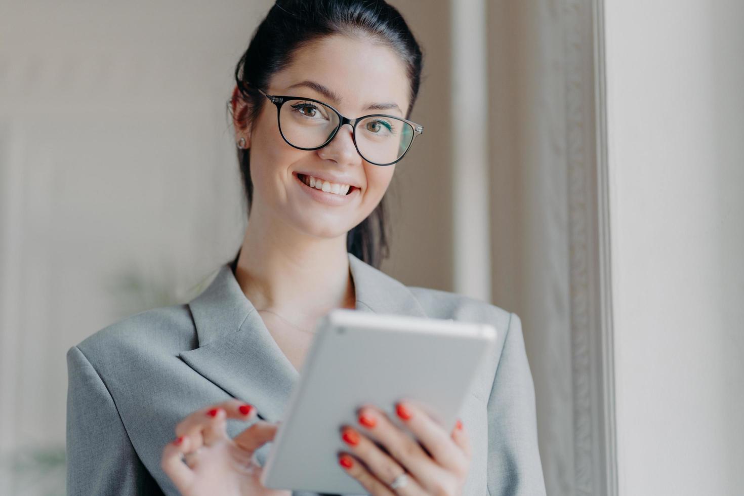 Indoor shot of happy brunette entrepreneur reads electronic book on modern digital tablet, reads financial news in internet during work break, wears optical glasses, grey costume, poses indoor photo