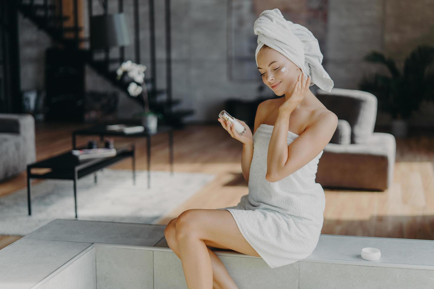 Smiling glad young woman touches healthy glowing skin, does daily beauty procedures at home, holds bottle of lotion, applies face cream, wrapped in bath towel, poses against cozy home interior photo