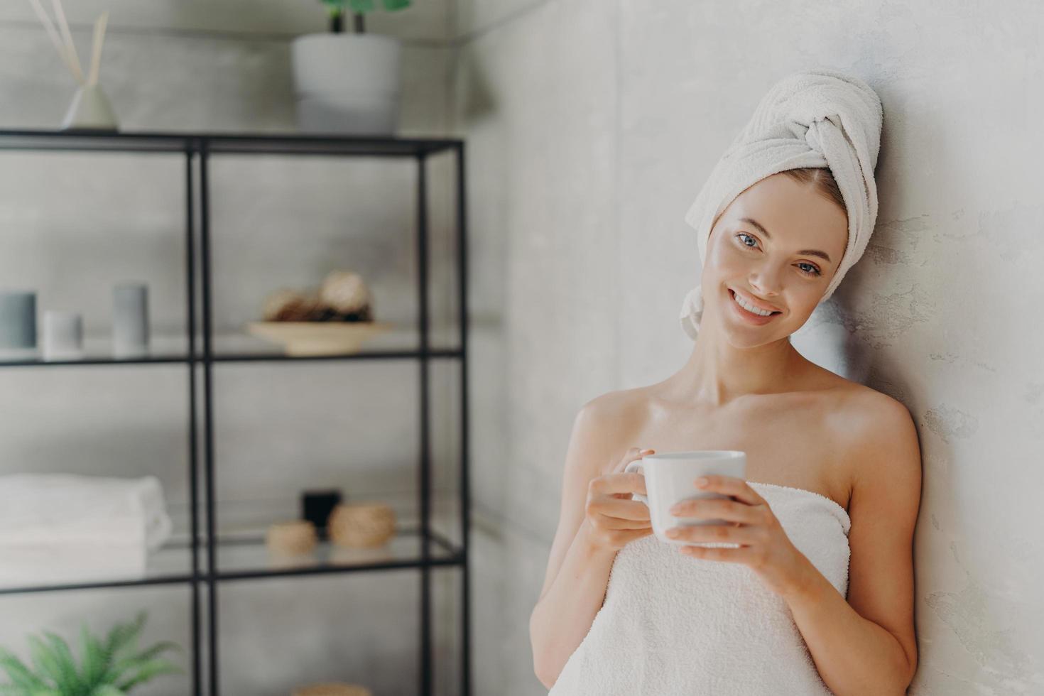 una mujer muy sonriente con una piel sana envuelta en una toalla blanca, se alegra en el interior, bebe café, tiene una expresión facial positiva, posa en el interior. personas, spa, mimos, cuidado de la piel y concepto de bienestar foto