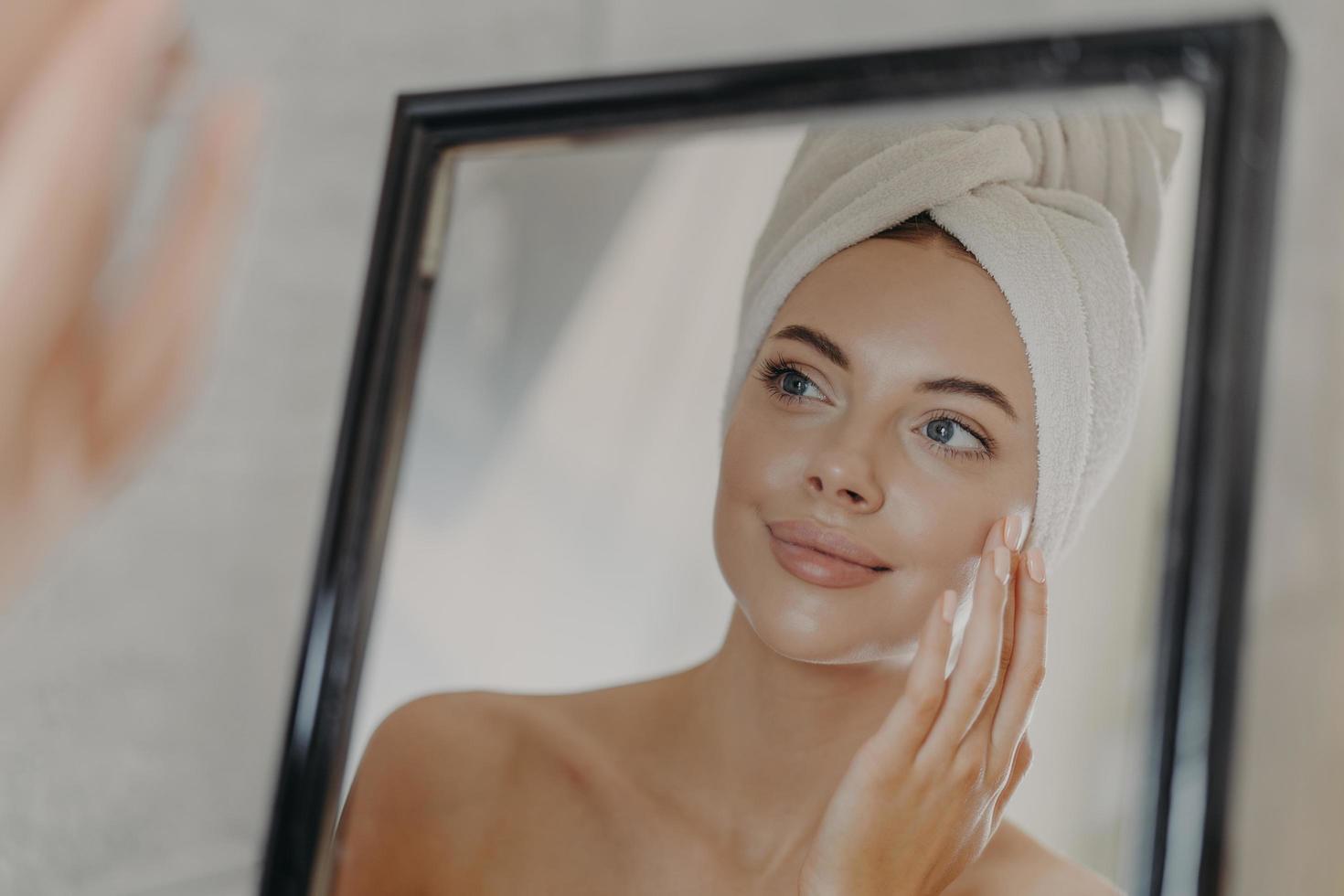 Young naked European woman touches soft skin on face, looks at herself in bathroom mirror, wears wrapped white towel on head after taking bath, poses bare shoulders. Beauty and skin care concept photo