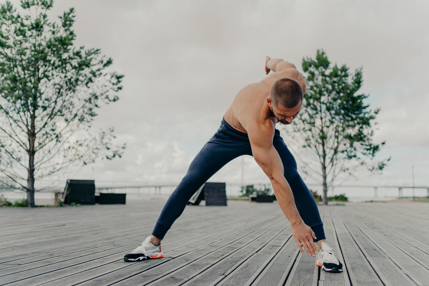 Muscular athlete does sport exercises, warms up body before cardio training, poses outdoor, dressed in active wear, leads active lifestyle. Sport and fitness concept. Bodybuilder with perfect biceps photo