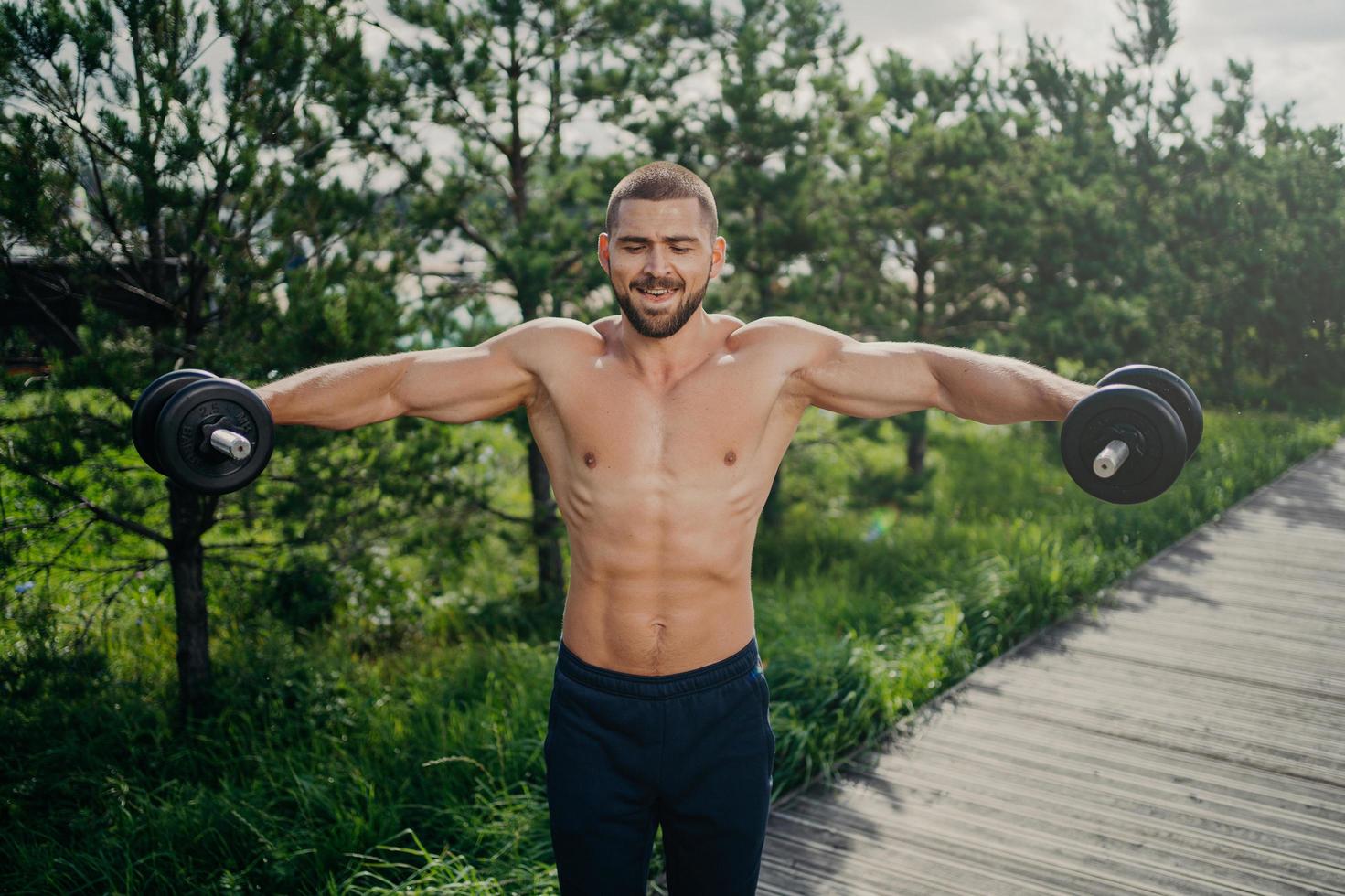 Fit adult man lifts heavy weight during workout session outdoor, poses with naked torso, has well shaped muscular body, demonstrates his power, lifts barbells. Motivated bodybuilder at street photo