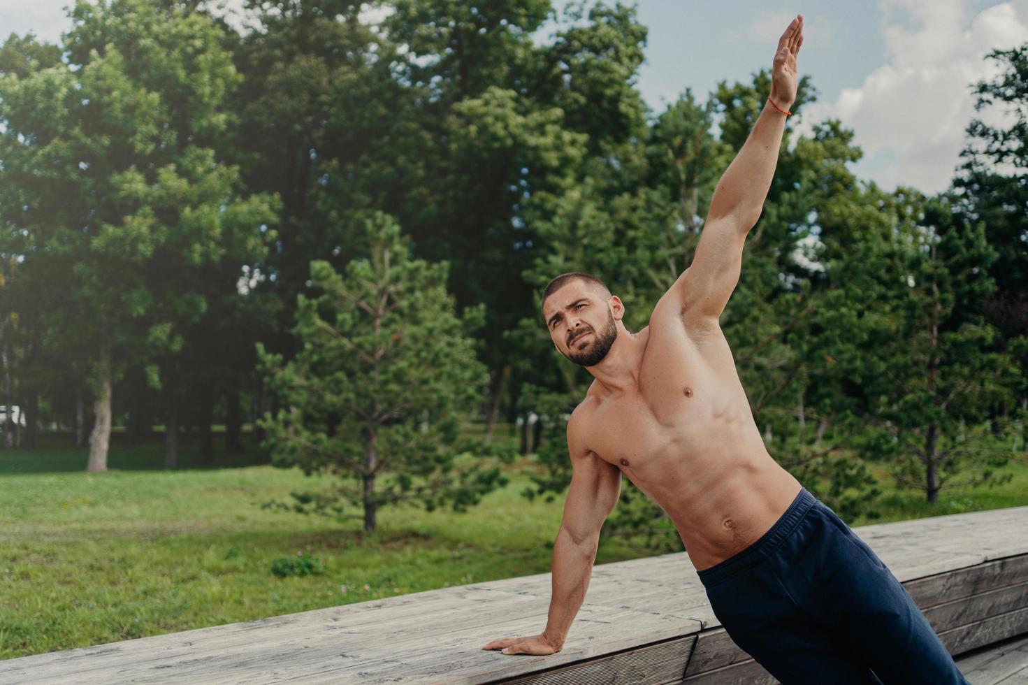 Strong young man stands in side plank on one arm, finds balance, poses at park near trees, practices yoga outdoor, leads active healthy lifestyle, has muscualr strong body. Motivated bodybuilder photo