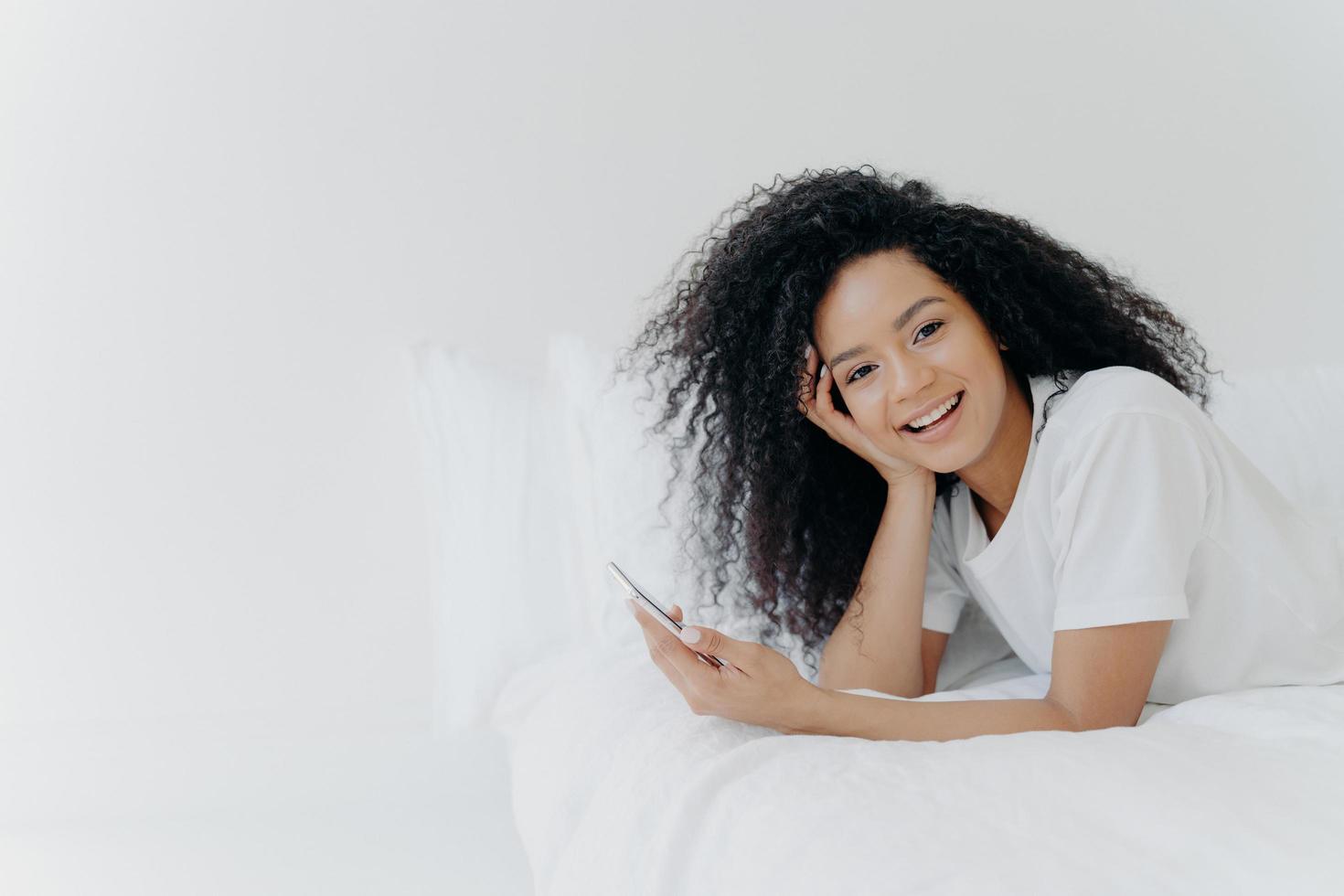 Overjoyed relaxed curly young African American woman enjoys spare time at home, lies in bed with modern cellphone, uses phone in bedroom, sends text message to lover, has lazy morning, good time photo