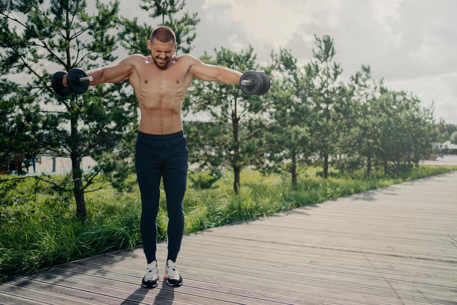 Photo of athletic strong fitness man works out with barbells, demonstrates power and energy, leads active lifestyle, shows strength, motivation, does biceps lifting, has physical workout outdoor