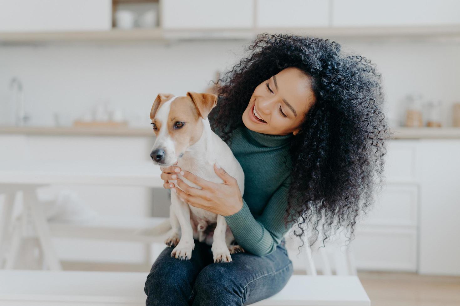 Happy female owner of jack russell terrier dog, feels responsibility of caring about pet, has bushy dark curly hair, sits against blurred kitchen background. People and relationship with animals photo