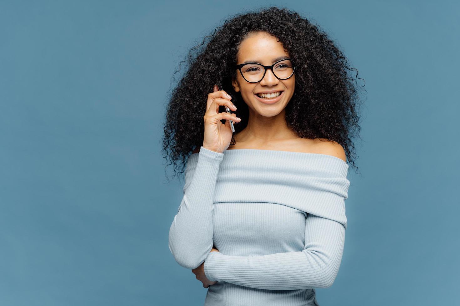 Indoor shot of positive lovely woman has telephone conversation, keeps mobile phone near ear, focused aside, wears casual jumper, isolated on blue background. People, technology, communication photo