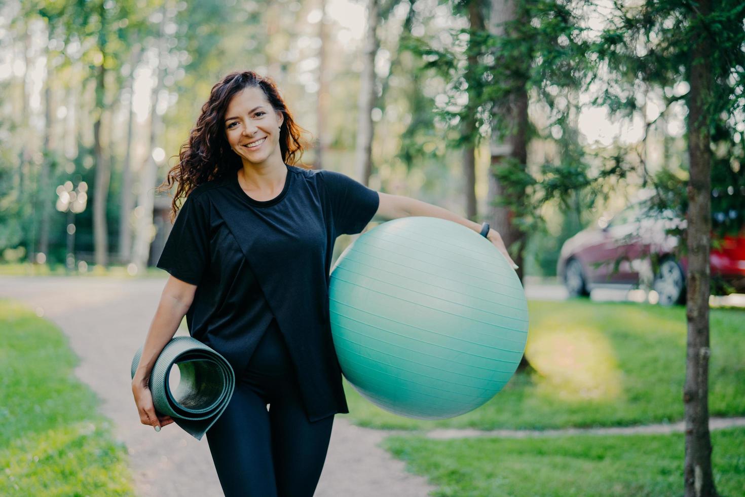 Pleased brunette woman dressed in black t shirt, holds rolled up karemat and fitness ball, prepares for aerobic exercises, smiles pleasantly, poses against nature background. People and sport concept photo