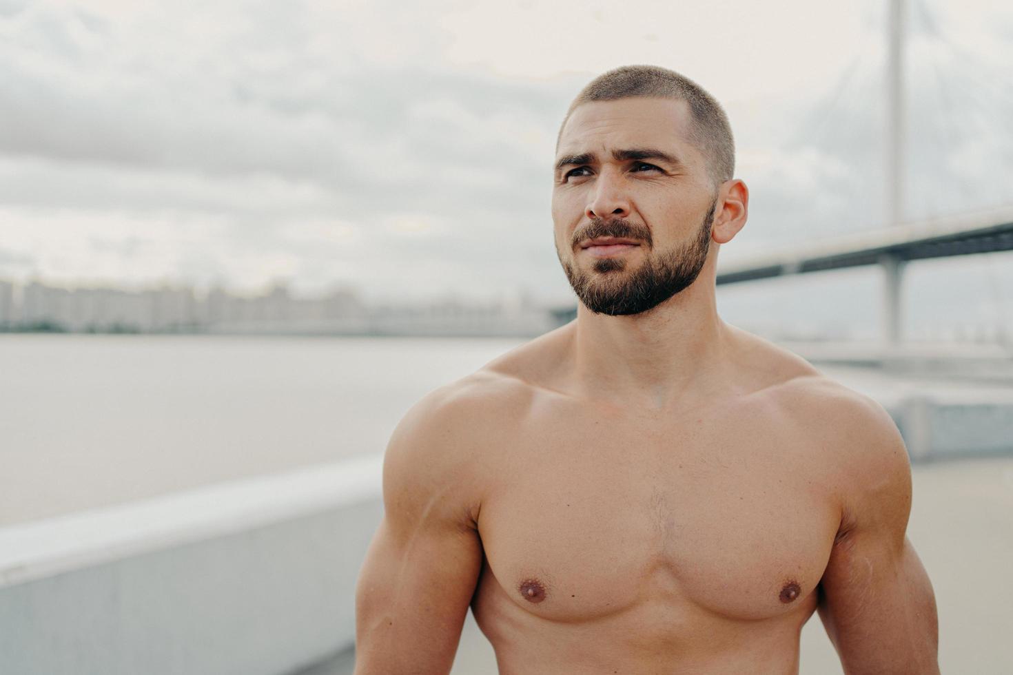 Horizontal shot of muscular man with naked torso thick beard looks somewhere into distance, relaxes outdoors, stands against river background, takes break after jogging, has athletic body shape photo