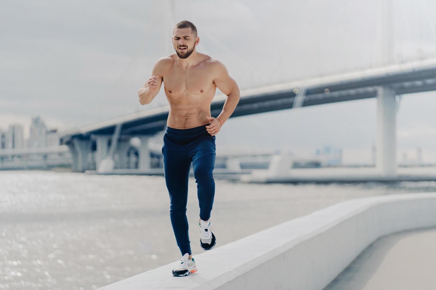 Athletic young bearded man goes in for sport has morning run breathes deeply prepares for running marathon. Motivated sportsman jogging by river has strong healthy body. Fitness and sport concept photo