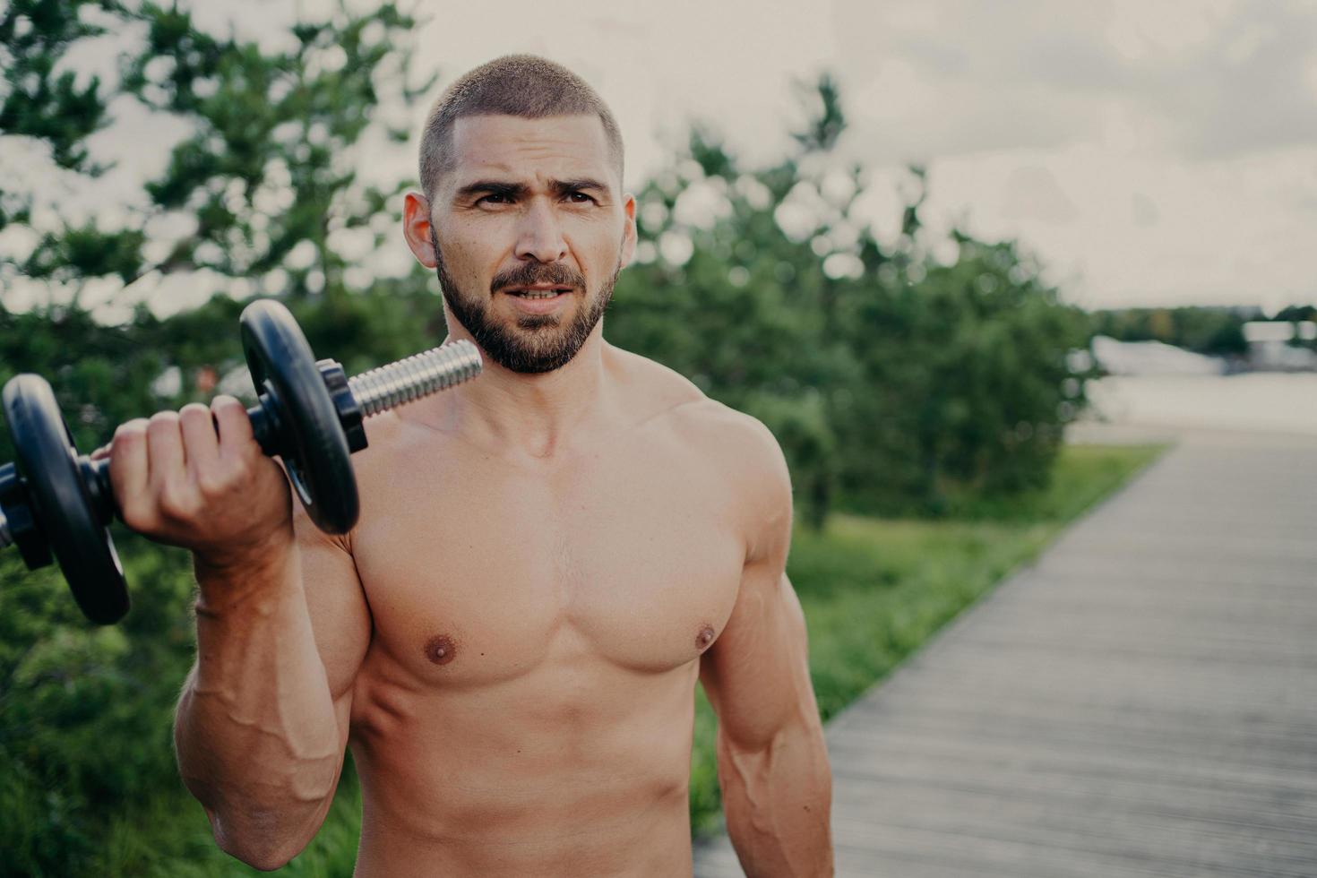 Horizontal shot of handsome muscular man lifts barbell outdoor, has athletic torso, gets ready for weight lifting training, has muscular arms, poses against street background. Developing strength photo