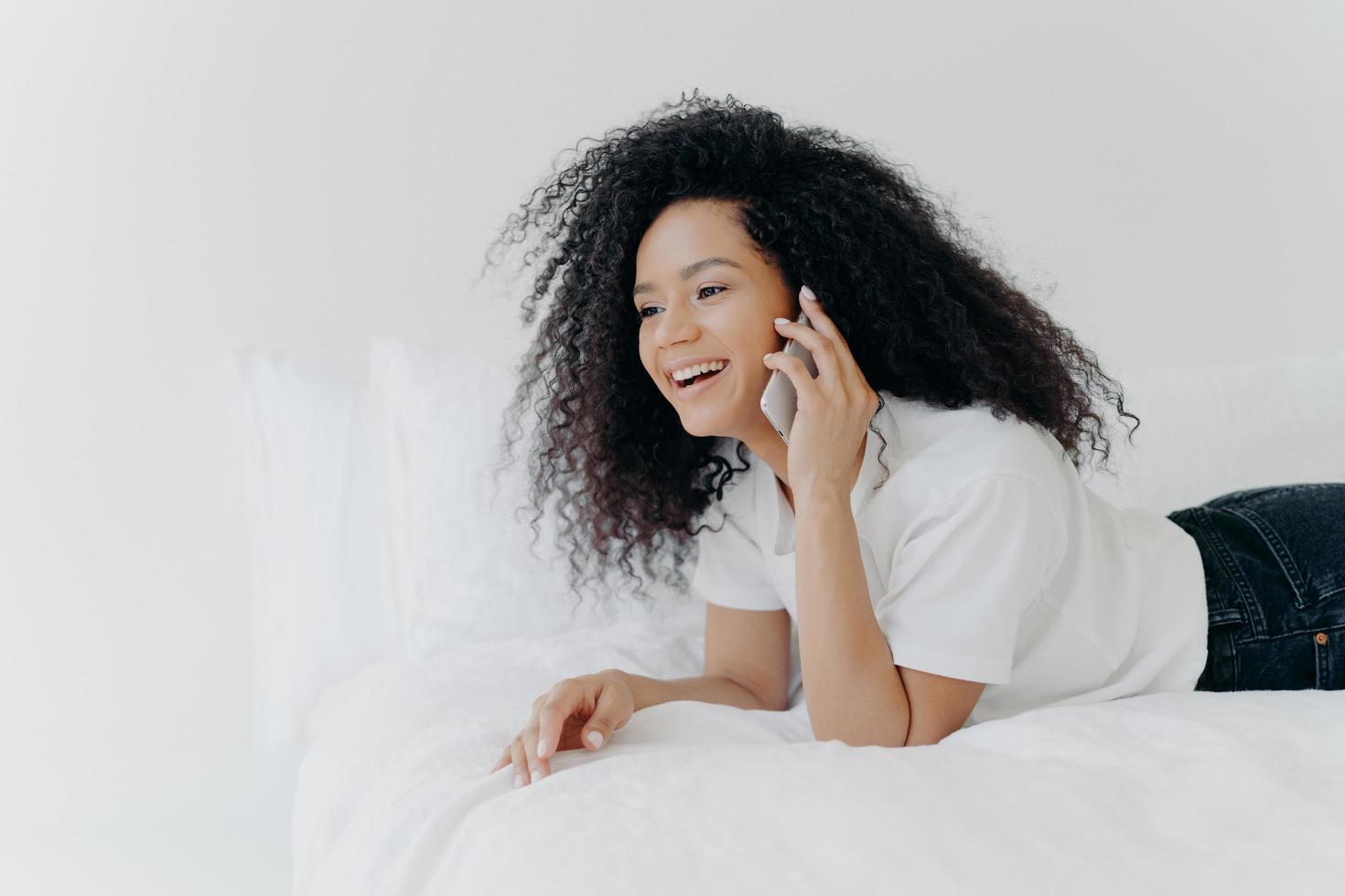 Encantadora mujer joven con cabello rizado, habla con su novio a través de un teléfono inteligente, descansa en la cama por la mañana, se ríe de una broma graciosa, se viste informalmente, está en casa contra una pared blanca. concepto de comunicación foto