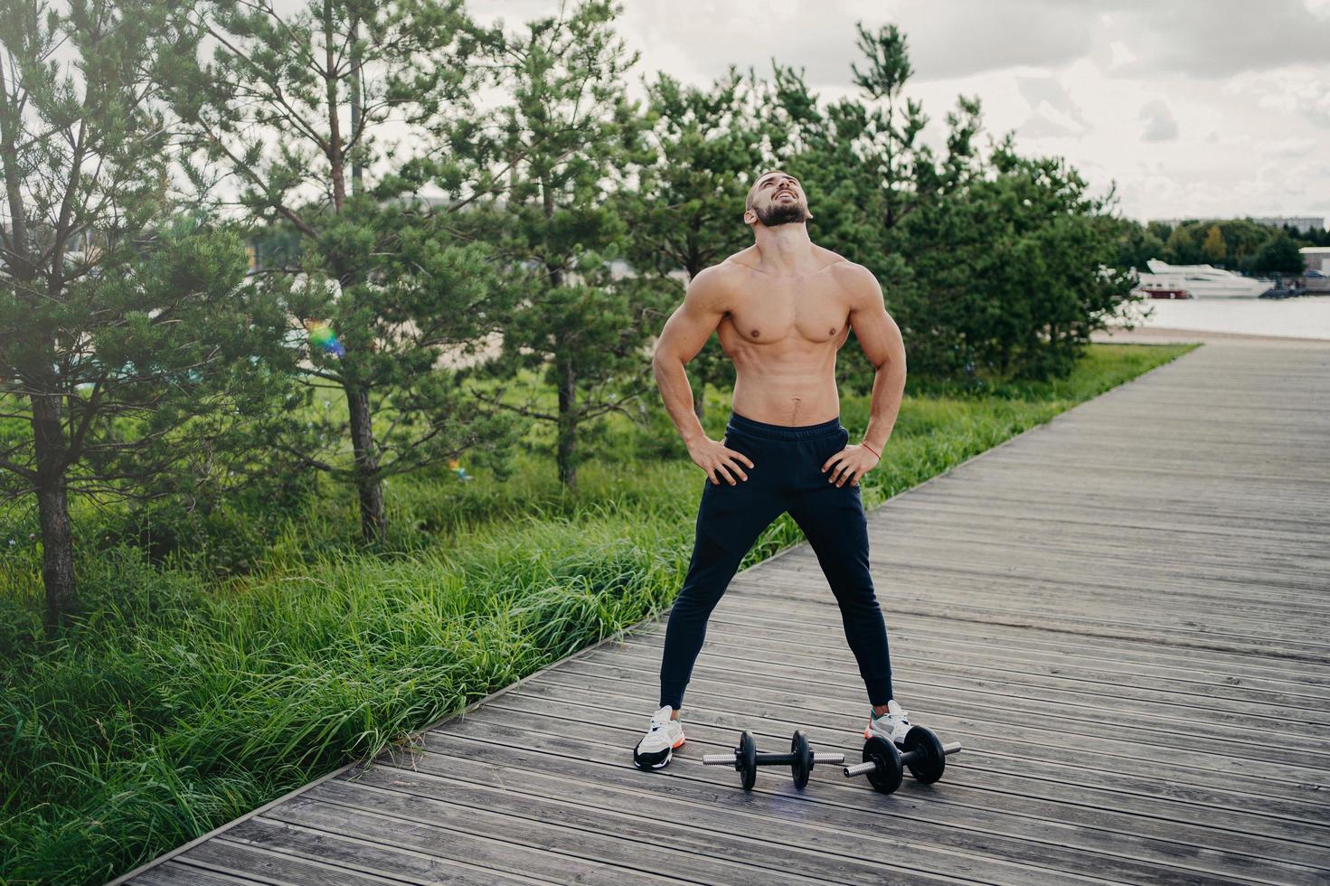 Healthy muscular man stands with naked torso, raises head above and breathes deeply, wears sport clothes, has workout with barbells, poses outdoor. Bodybuilding, active lifestyle, fitness concept photo
