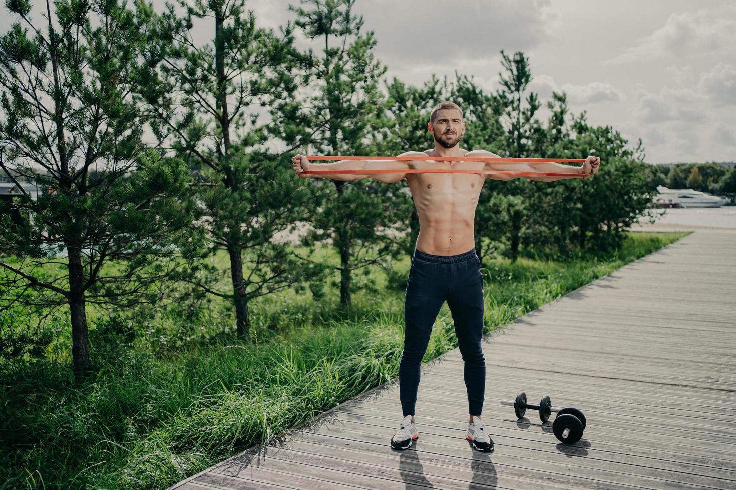 Workout, healthy lifestyle and bodybuilding concept. Strong muscular European man stretches resistance band, wears sneakers, stands outdoor, enjoys exercising in open air, has athletic body. photo