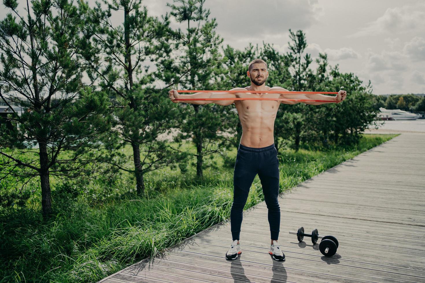 entrenamiento, estilo de vida saludable y concepto de culturismo. un hombre europeo fuerte y musculoso estira la banda de resistencia, usa zapatillas de deporte, se para al aire libre, disfruta hacer ejercicio al aire libre, tiene un cuerpo atlético. foto