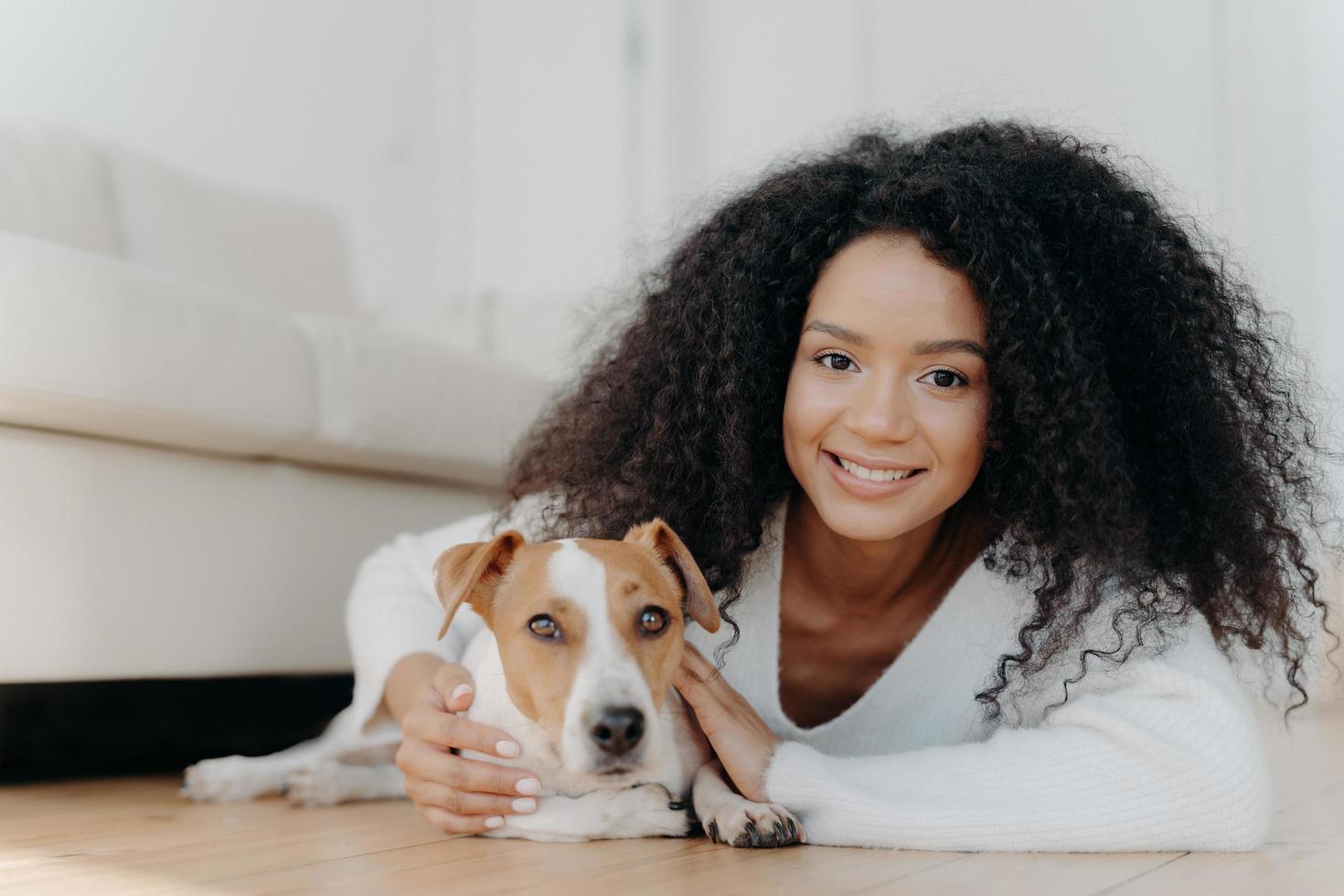 Pretty girl with Afro hair, lies on floor with dog, expresses pleasant emotions, poses in living room near couch, bought pet in new apartment. Woman host with beloved animal at home, share good moment photo