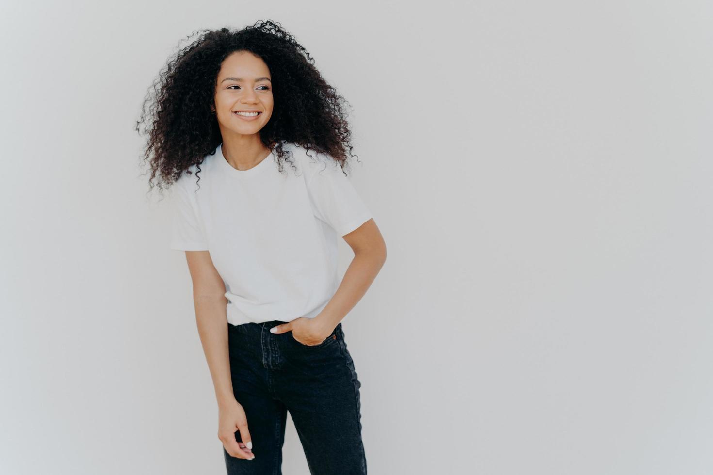 Young curly haired lady has slim figure, wears white t shirt and jeans, looks aside with happy expression, shows healthy teeth, notices funny scene on right side, isolated on white background photo
