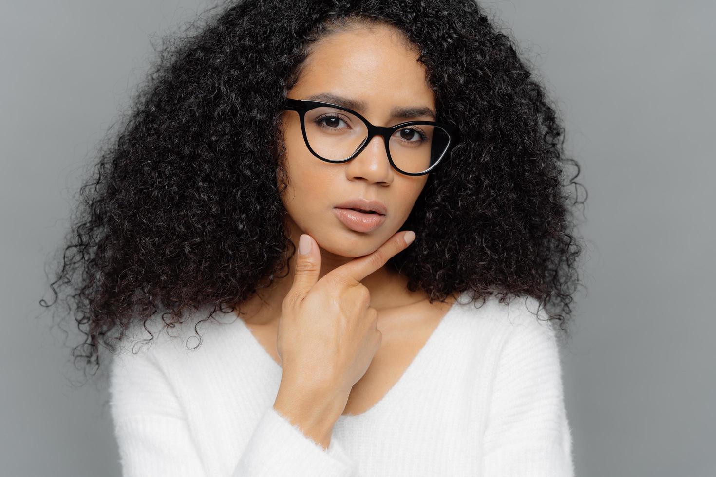 Close up portrait of thoughtful African American woman holds chin, looks mysteriously at camera, has bushy crisp hair, wears optical glasses and white jumper, makes plan, isolated over grey background photo