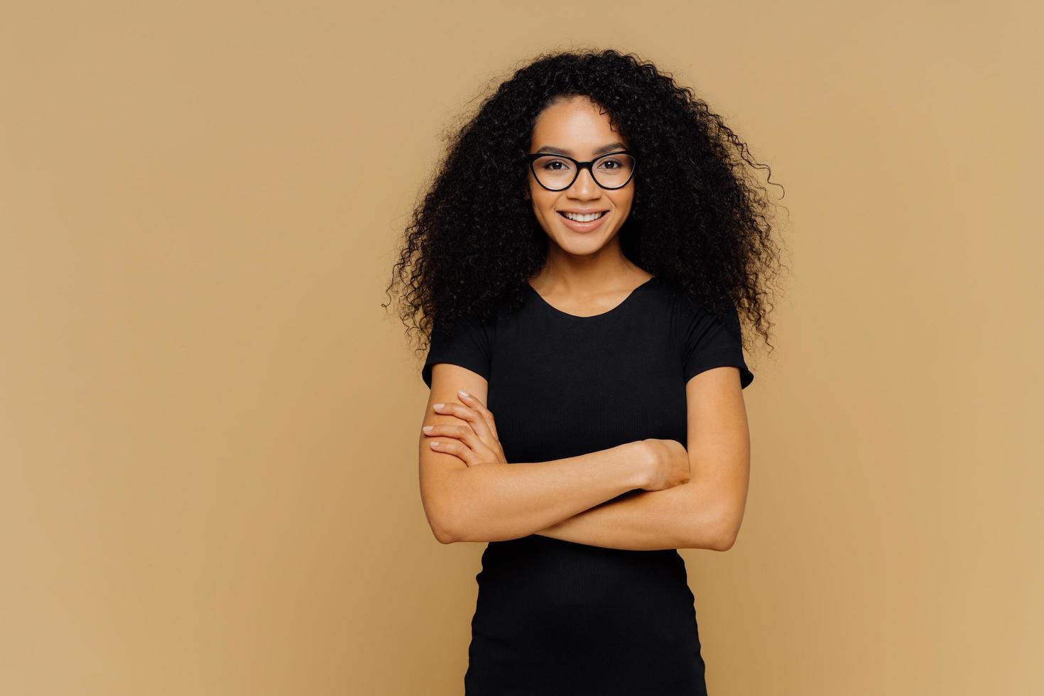 Slim satisfied woman with Afro haircut, wears black casual clothes, optical glasses, has confident expression, listens interlocutor, isolated on beige background. Cute teenage girl enjoys life. photo