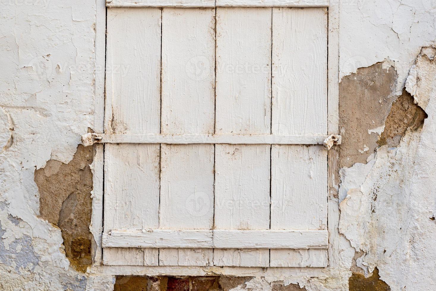 White cracked wall with a window closed with a painted white wooden shutters and locked with two locks photo