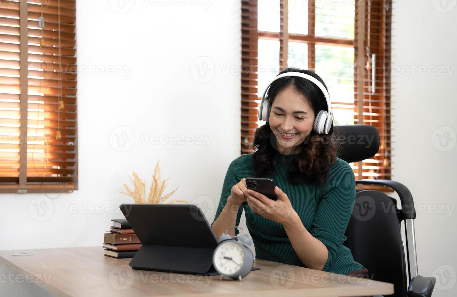 Asian woman school student wear headphones hold smartphone using distance learning mobile app online watching video course or zoom calling making notes in workbook sit at home. photo