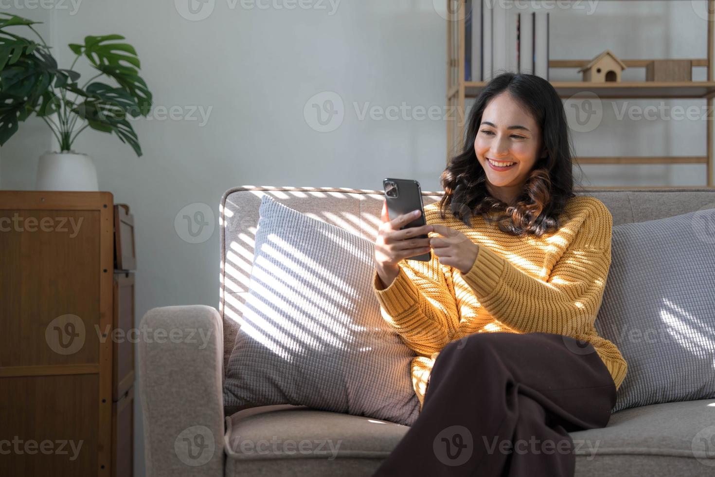 Happy girl checking social media holding smartphone at home. Smiling young latin woman using mobile phone app playing game, shopping online, ordering delivery relax on sofa. photo