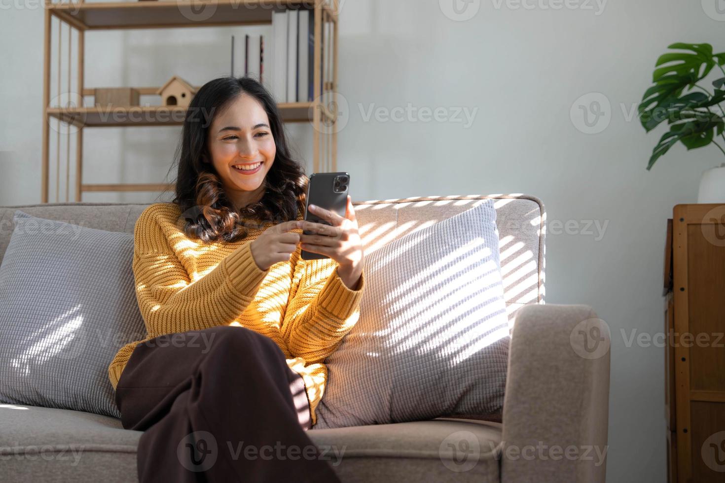 Happy girl checking social media holding smartphone at home. Smiling young latin woman using mobile phone app playing game, shopping online, ordering delivery relax on sofa. photo