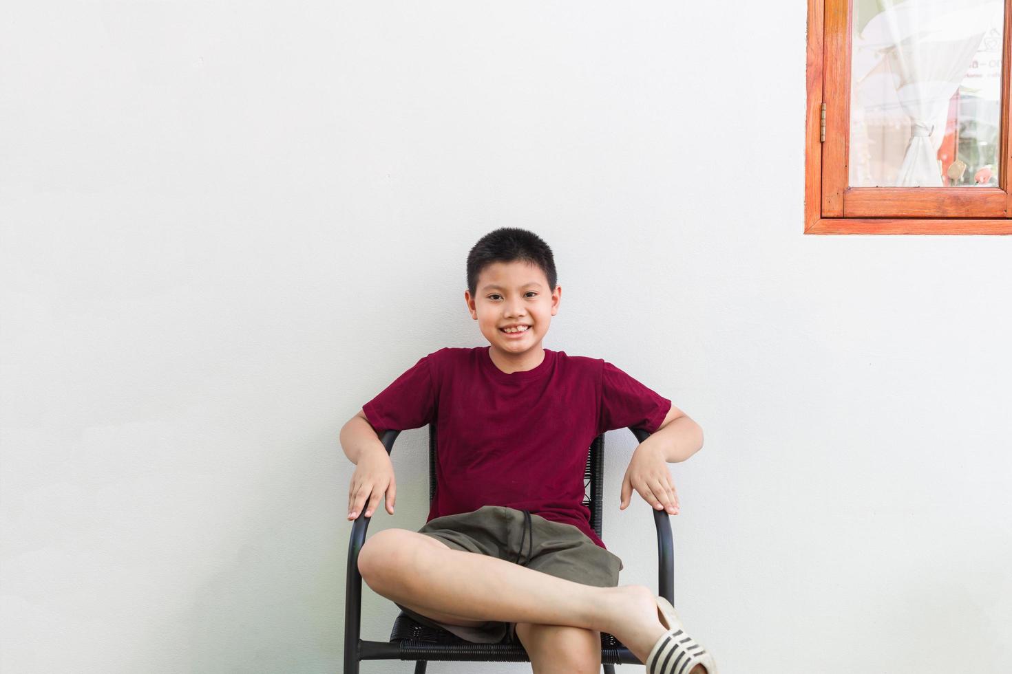 niño asiático tailandés sentado en una silla sonriendo, luciendo guapo y divertido en la pared blanca de un café en vacaciones foto
