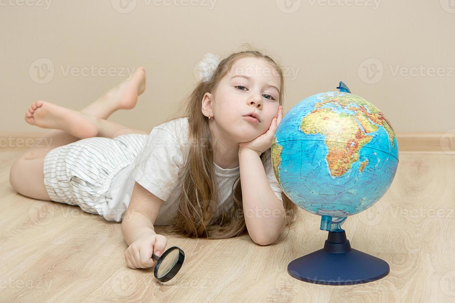 pensive little girl lies on the floor next to a globe, holds a magnifying glass in one hand, put the other under her head and argues. The concept of a smart child, education, plans for the future photo