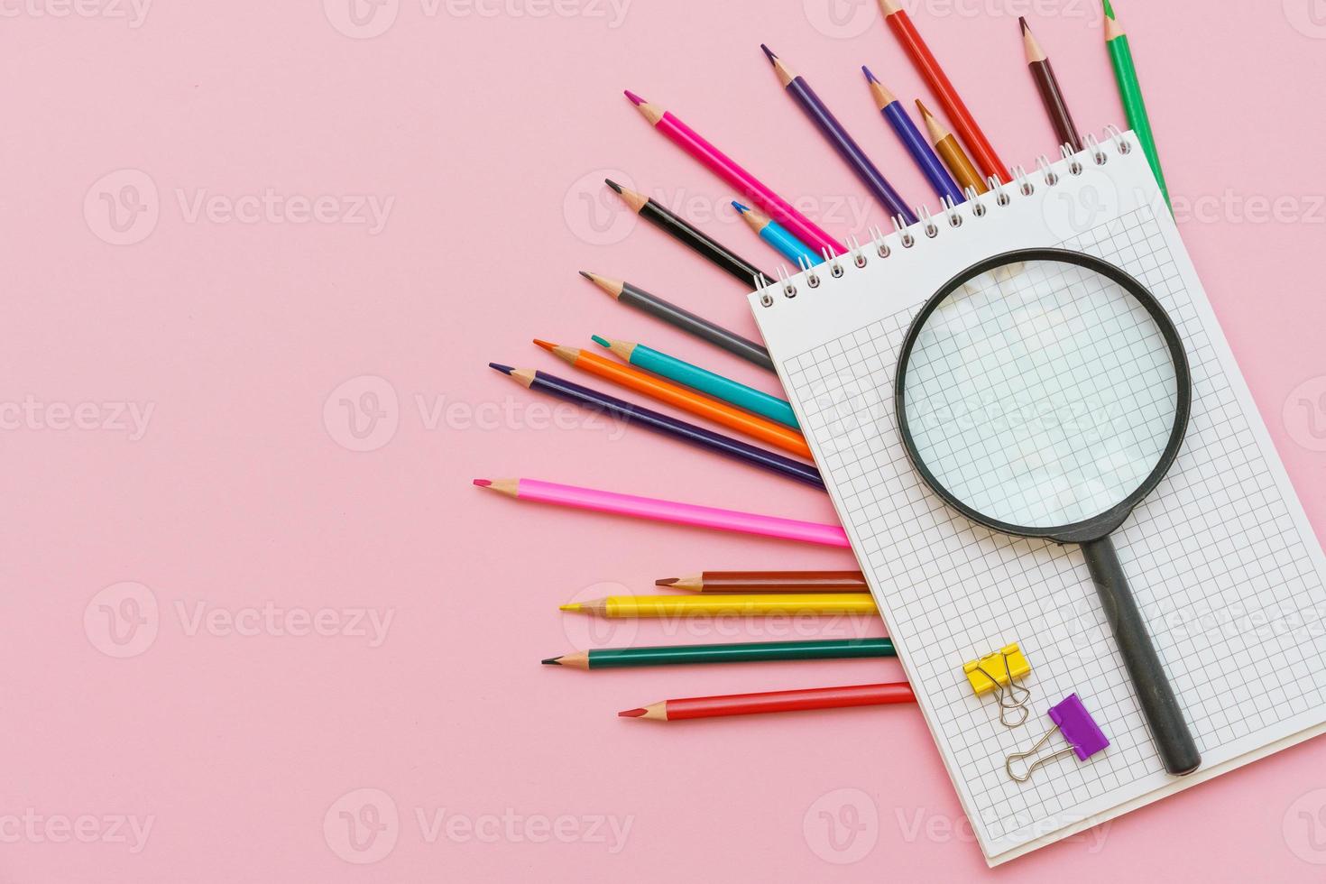 Magnifying glass, pencils and a blank white notepad on a spring on a pink photo
