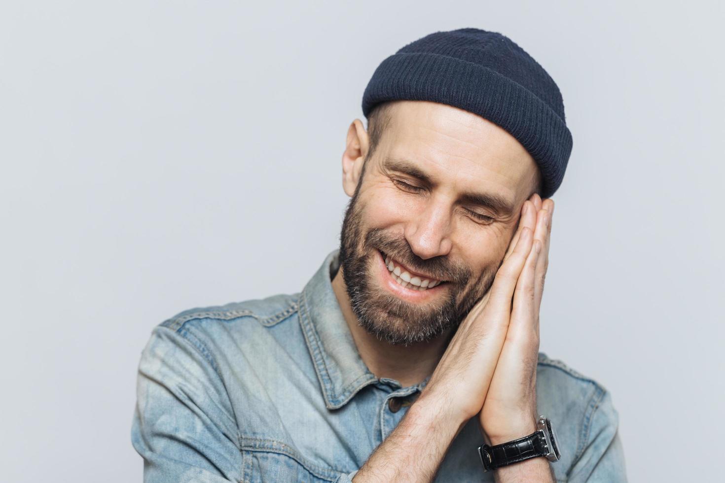 Positive bearded male pretends sleeping, has pleasant dreams, rests on hands, smiles joyfully, wears black stylish hat, isolated over white background. People, good rest and emotions concept. photo