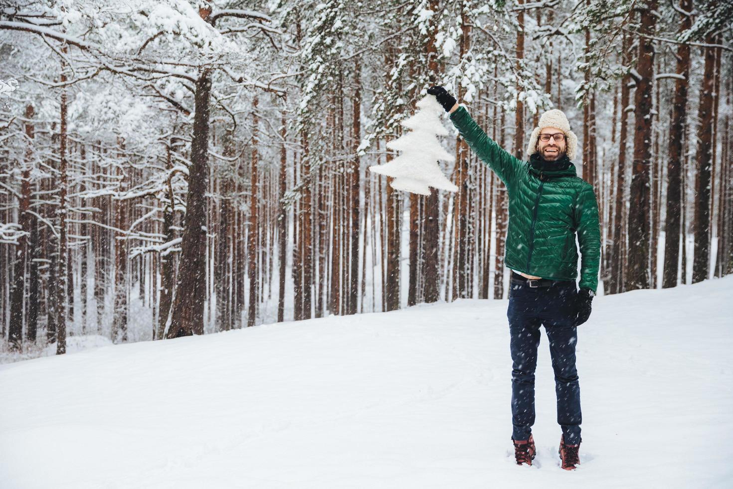 Good looking middle aged male model wears warm fur cap with ear flaps and green down padded coat, holds white artificial fir tree, stands on ground covered with snow, has happy expression photo