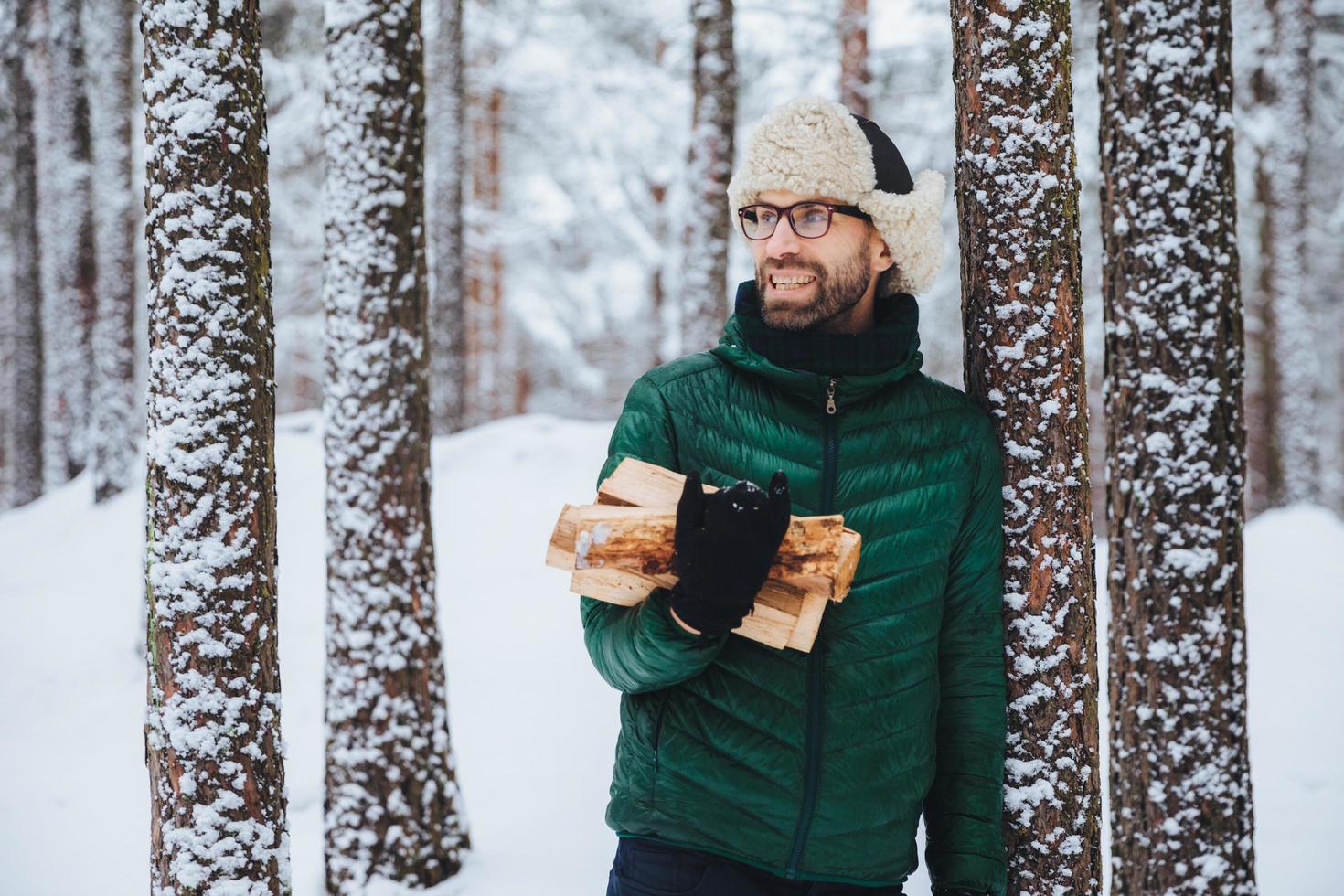 Happy bearded man in green jacket, stands near tree, holds firewood, looks thoughtfully aside, has cheerful expression, stands against snowy forest background. Fashionable pensive male outdoor photo