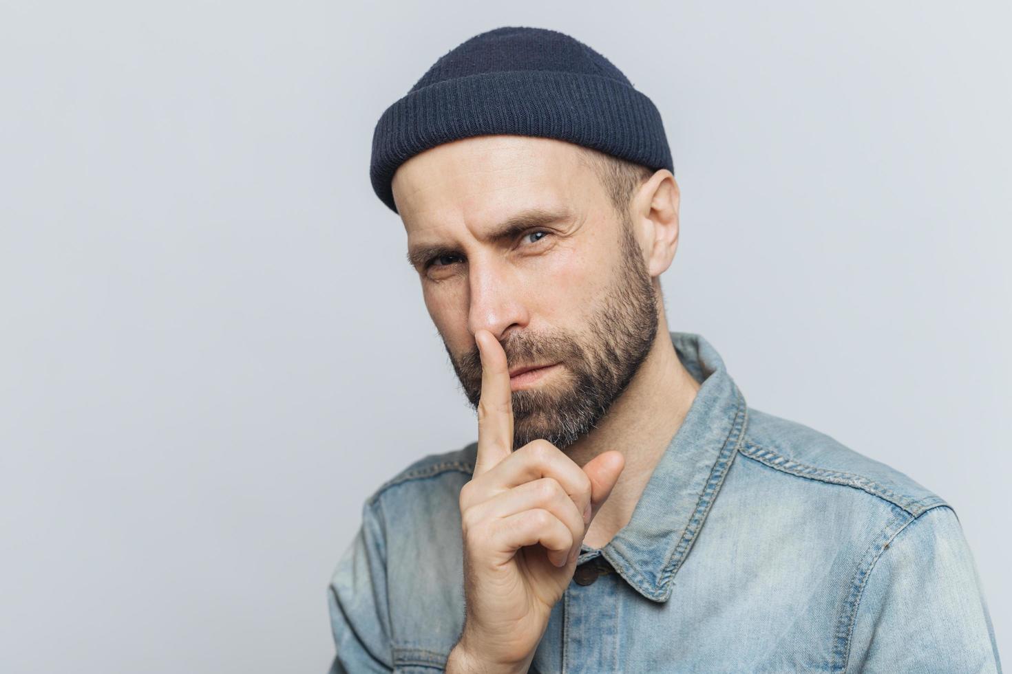 Portrait of serious bearded male with attractive look, keeps fore finger on lips, looks with secret expression, asks to keep silence and not tell his secret to anybody, isolated over grey background photo