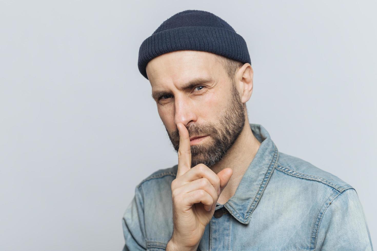 retrato de hombre barbudo serio con aspecto atractivo, mantiene el dedo índice en los labios, mira con expresión secreta, pide guardar silencio y no contar su secreto a nadie, aislado sobre fondo gris foto