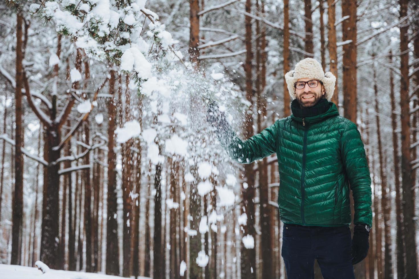 Smiling delightful male dressed in warm clothes, stands in winter forest, throws snow in air, has fun alone, has good mood, expresses positive emotions and feelings. Positiveness concept photo